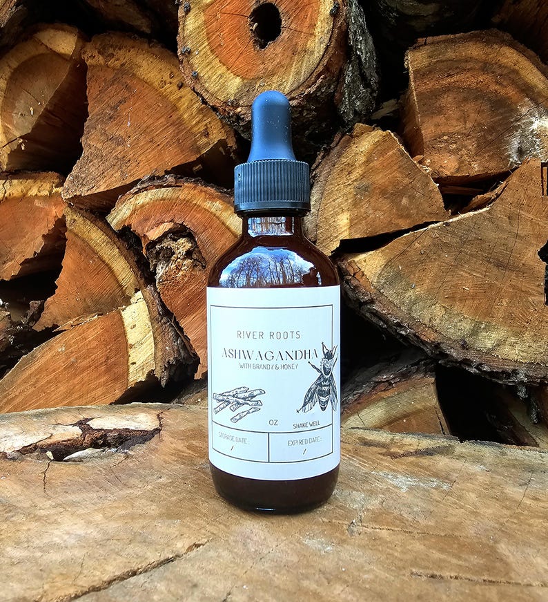 May include: A brown glass bottle with a blue dropper top. The label reads "River Roots Ashwagandha with Brandy & Honey." The label also features an illustration of a bee and plant roots. The bottle is sitting in front of a stack of wood.