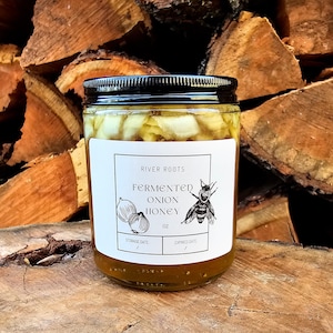 May include: A jar of fermented onion honey with a black lid and a white label. The label reads "River Roots Fermented Onion Honey" with illustrations of onions and a bee. The jar is filled with honey and onion pieces.