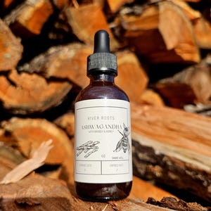 May include: A brown glass bottle with a black dropper, labeled "River Roots Ashwagandha with Brandy & Honey." The label features illustrations of herbs and a bee. The bottle is set against a backdrop of stacked firewood.