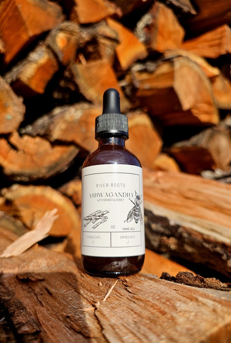May include: A brown glass bottle with a black dropper, labeled "River Roots Ashwagandha with Brandy & Honey." The label features illustrations of herbs and a bee. The bottle is set against a backdrop of stacked firewood.