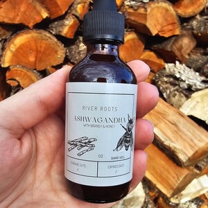 May include: A dark glass bottle with a black dropper, labeled "RIVER ROOTS ASHWAGANDHA WITH BRANDY & HONEY." The label features a bee illustration and text. The bottle is held in a hand against a backdrop of stacked firewood.