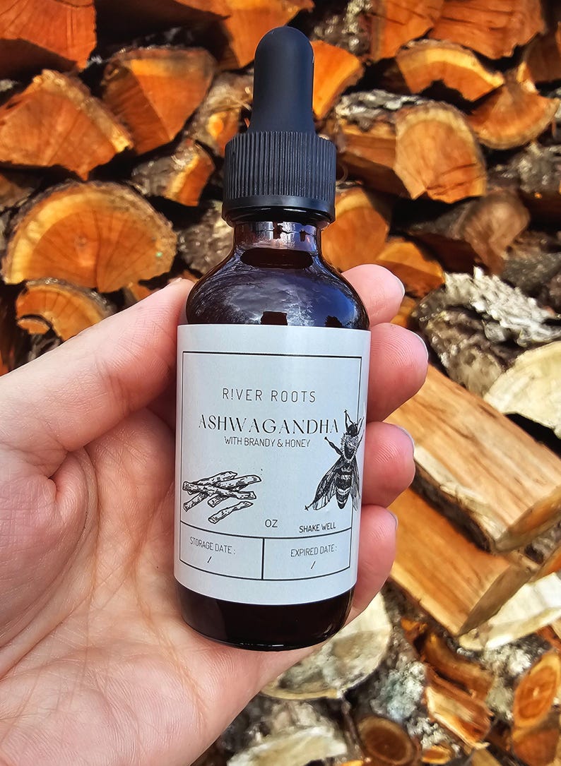 May include: A dark glass bottle with a black dropper, labeled "RIVER ROOTS ASHWAGANDHA WITH BRANDY & HONEY." The label features a bee illustration and text. The bottle is held in a hand against a backdrop of stacked firewood.