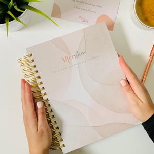 May include: A white and pink self-care planner and journal with the title "Afterglow" and a gold spiral binding.