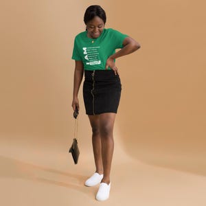 May include: A green t-shirt with white text featuring a list of names and the word "MAMA" in a stylized font. The person is wearing a black skirt with a gold zipper and white shoes. A black purse hangs from the person's hand.