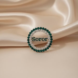May include: A circular brooch with a double row of gemstones. The outer row features emerald green stones, while the inner row has clear, sparkling stones. The word "Soror" is displayed in the center. The brooch is set against a satin background.