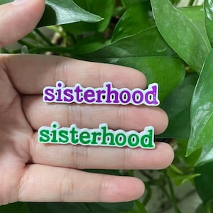 May include: Two white plastic pins with the word "sisterhood" written in green and purple letters. The pins are held in a hand.