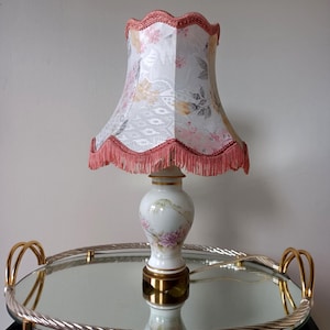 Könnte beinhalten: Eine Tischlampe mit einem weißen Keramikfuß, der mit gemalten rosa und violetten Blumenmotiven sowie goldenen Verzierungen versehen ist. Der gewellte Stoffschirm hat einen weißen Hintergrund, ein florales Muster und einen rosa Fransenbesatz.