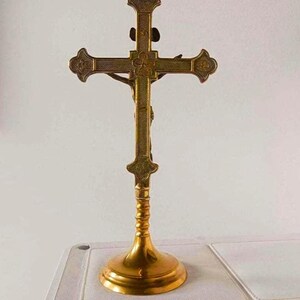 May include: A gold-toned metal crucifix standing on a circular base. The cross features a textured surface with decorative ends and a central floral motif. A figure is attached to the front of the cross, extending across the horizontal bar.