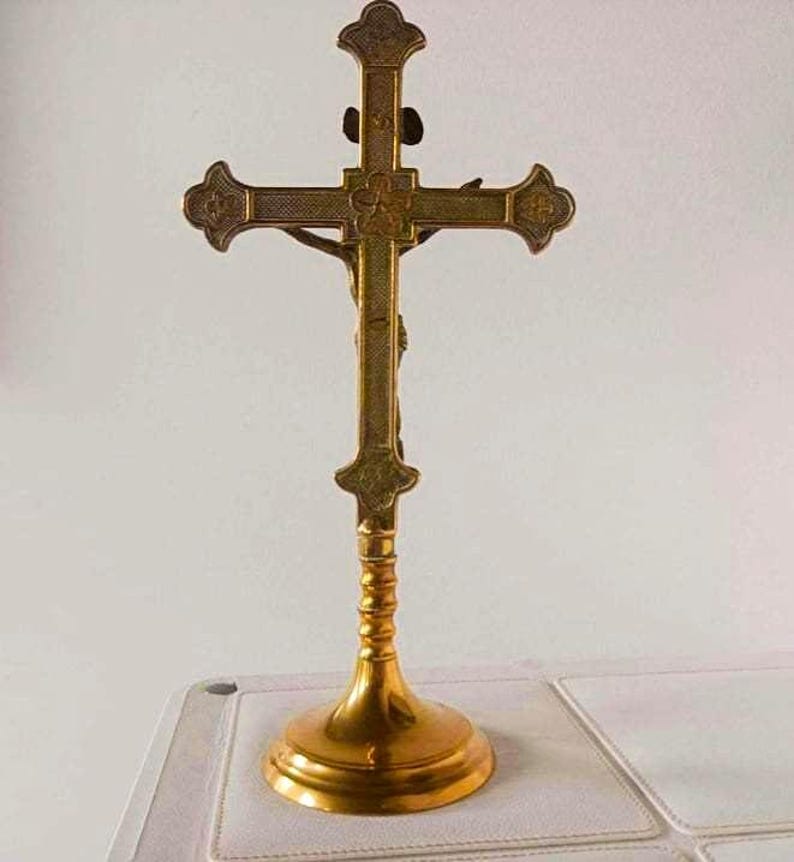 May include: A gold-toned metal crucifix standing on a circular base. The cross features a textured surface with decorative ends and a central floral motif. A figure is attached to the front of the cross, extending across the horizontal bar.