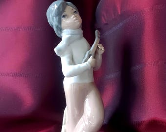 May include: A glazed porcelain figurine of a person with gray hair wearing a white turtleneck sweater and light brown pants. The figure is standing and holding a small musical instrument, possibly a ukulele, with gray shoes and a glossy finish.