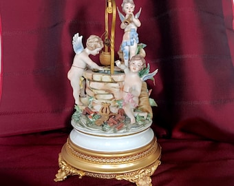 May include: A porcelain figurine featuring three winged cherubs surrounding a stone-textured well. The well includes a gold-toned metal crane and bucket. The base is white with a gold-toned, ornate pedestal stand featuring four decorative feet.