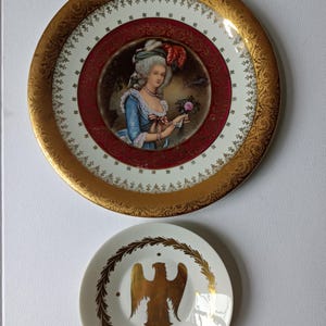 May include: A large decorative plate featuring a central portrait of a person in 18th-century attire with a gold-patterned rim and a deep red border, accompanied by a smaller white saucer displaying a gold eagle emblem encircled by a laurel wreath.