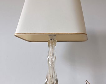 May include: A table lamp featuring a clear glass base with a twisted, spiral design. The lamp is topped with a rectangular, off-white fabric lampshade that has a thin, gold-colored trim along the top and bottom edges. A brown power cord is attached.