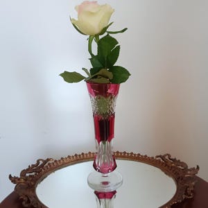 Puede incluir: Un florero alto y esbelto de vidrio tallado en color rojo rubí y transparente, con un patrón de diamantes en la parte superior y una base lisa y acampanada. Una rosa color crema con hojas verdes se encuentra dentro del florero sobre una bandeja dorada.