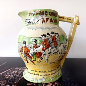 May include: A vintage ceramic pitcher with a hand-painted scene of a fair, featuring figures on horseback and the text "Widdicombe Fair." The pitcher has a cream-colored base with a green rim and handle, and is decorated with colorful illustrations.
