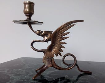 May include: A brass candlestick holder shaped like a mythical winged creature with a long, curved neck and a coiled tail. The candle cup sits atop a flat, scalloped base supported by the creature's head, featuring detailed wing feathers and metal texture.