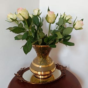 May include: A bouquet of cream and pale pink roses with green leaves arranged in a gold-rimmed glass vase. The vase features a scalloped gold pattern and sits on an oval, ornate gold-toned metal tray with a mirrored base.