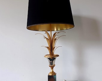 May include: A table lamp featuring a black drum shade with a gold interior lining. The base is constructed of gold-toned metal shaped like a stylized plant or pineapple, set atop a square metallic pedestal with a reflective, dark-colored block.