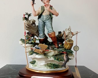 May include: A porcelain figurine of a fisherman standing on a rocky base with a fishing rod, a basket, and a caught fish. The figure wears a hat, vest, and trousers. A metal plaque on the wooden base reads PORCELLANE Il Pescatore.