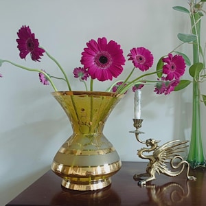 May include: A yellow glass vase with a flared rim and gold bands, containing several magenta gerbera daisies. Beside it sits a brass dragon-shaped candle holder with a white taper candle, and a tall, slender green glass vase with foliage.