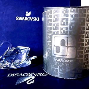 Puede incluir: Una figura de cisne de cristal facetado transparente colocada junto a una caja cilíndrica plateada con un patrón de logotipo repetitivo y el texto Swarovski Silver Crystal, frente a una caja azul oscuro con el logo del cisne y nombre Swarovski.
