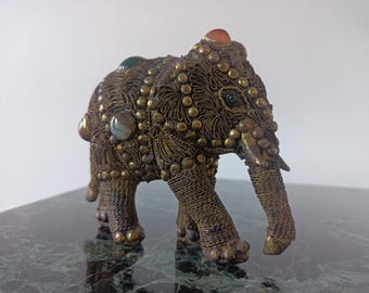 May include: A decorative elephant figurine crafted from dark metallic wire filigree. The surface is adorned with rows of small brass-colored studs and several smooth, polished cabochon stones in shades of green, orange, and translucent white.
