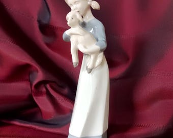May include: A porcelain figurine of a person with light hair styled in pigtails, wearing a long white dress with a grey bodice and hem, holding a small white lamb with grey hooves against their chest in a gentle embrace.
