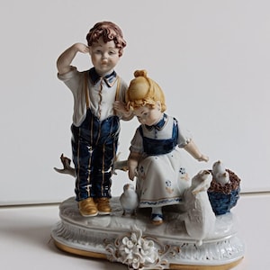 Puede incluir: Una figura de porcelana que representa a dos niños con pájaros. El niño lleva una camisa blanca, pantalones azules y zapatos dorados. La niña viste un vestido blanco con detalles azules. También hay un conejo blanco y una cesta azul con pájaros.