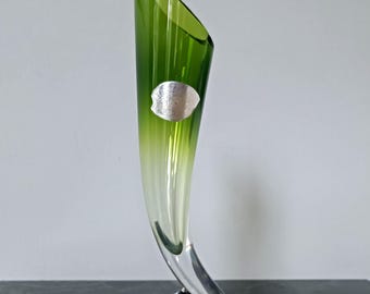 May include: A tall, horn-shaped glass vase featuring a vibrant green gradient that transitions into a clear, solid glass base. The upper rim is angled, and a small, circular silver-colored sticker is affixed to the front of the green glass body.