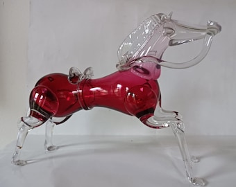 May include: A glass horse figurine featuring a deep red body and clear glass legs, mane, and head. The horse is depicted in a galloping pose with a small clear glass saddle detail on its back and a curved neck, crafted from smooth, translucent glass.