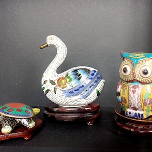 Op de afbeelding: Drie cloisonné dierenbeeldjes op houten standaards: een zwarte en rode schildpad, een witte zwaan met blauwe en florale accenten, en een veelkleurige uil met turquoise, gele en paarse patronen. Elk stuk is voorzien van gedetailleerd draadwerk.