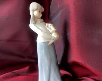 May include: A porcelain figurine of a person with light brown hair wearing a white long-sleeved top and a long, light blue skirt. The person is holding a small, light brown and white puppy close to their chest, with a smooth, glossy finish.