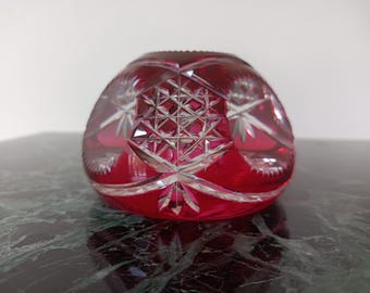 May include: A deep red cut glass bowl featuring intricate geometric patterns and starburst motifs. The glass has a thick, rounded shape with clear, faceted edges that contrast against the vibrant crimson color of the material.