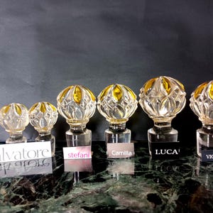 May include: Six clear glass decanter stoppers with faceted spherical tops and amber-colored accents. Each stopper is paired with a small label featuring the names Salvatore, stefani, Camilla, LUCA, and VICENZA, arranged in a row on a dark surface.
