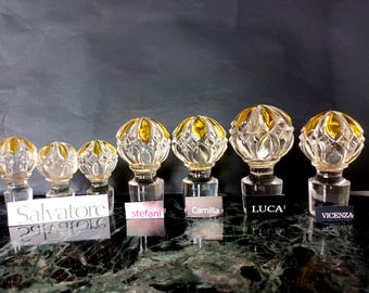 May include: Six clear glass decanter stoppers with faceted spherical tops and amber-colored accents. Each stopper is paired with a small label featuring the names Salvatore, stefani, Camilla, LUCA, and VICENZA, arranged in a row on a dark surface.