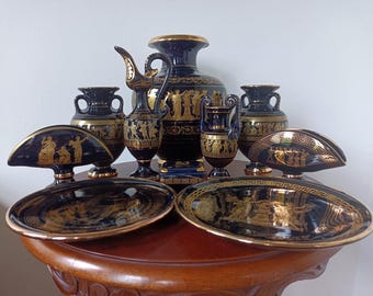 May include: A collection of dark blue ceramic decorative items featuring gold-colored Greek-style figures and geometric patterns. The set includes a large vase, a pitcher, two smaller vases, two plates, and two semi-circular stands with gold trim.