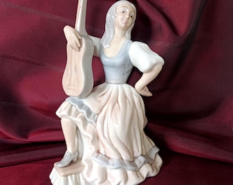 May include: A porcelain figurine of a person with long hair wearing a light blue bodice and a flowing, ruffled skirt. The figure is posed in a seated position, holding a light-colored guitar upright with one hand while resting the other on a hip.
