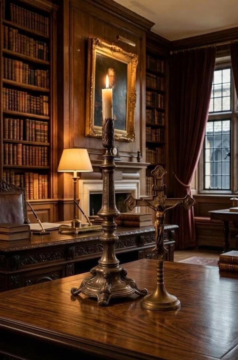 May include: A tall, ornate bronze candlestick holding a lit white candle, positioned next to a bronze crucifix on a polished wooden desk. The desk is set within a library featuring dark wood paneling, bookshelves, and a framed portrait.