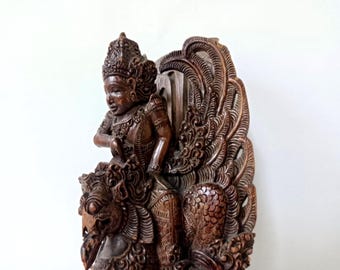 May include: A dark brown wooden sculpture depicting a figure wearing an ornate headdress and jewelry, seated atop a mythical creature with a bird-like head and large, intricately carved wings that extend upward behind the central figure.