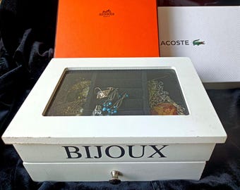 May include: A white wooden jewelry box with a glass lid and a single drawer featuring the word BIJOUX printed in black on the front. The interior is lined with black velvet and contains various pieces of jewelry, including earrings and chains.