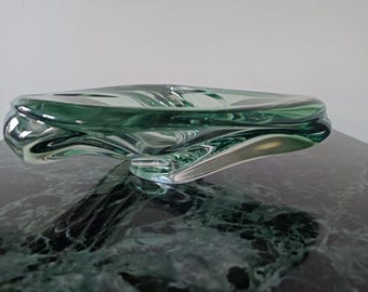 May include: A translucent, light green glass bowl featuring an organic, asymmetrical shape with fluid, undulating edges. The bowl has a thick, clear glass base and a smooth, polished surface that reflects light, creating depth and visual interest.