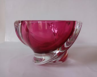 May include: A deep magenta glass bowl featuring a thick, clear glass base with a swirling, sculptural design. The bowl has a wide, open top and a smooth, polished finish, with the vibrant pink color concentrated in the center and fading into clear glass.