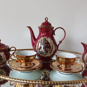 May include: A vintage tea set with a burgundy and gold color scheme. The set includes a teapot, sugar bowl, creamer, and two teacups with saucers. Each piece features a detailed illustration and gold trim.
