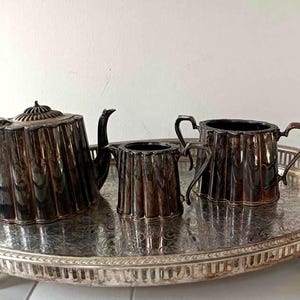 Op de afbeelding: Een verzilverd theeservies bestaande uit een theepot, een melkkan en een suikerpot met een geribbeld ontwerp, geplaatst op een ovaal dienblad met een sierlijk gegraveerd patroon en een opengewerkte rand, met een aangeslagen metalen afwerking.