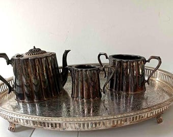 May include: A silver-plated tea set consisting of a teapot, a creamer, and a sugar bowl with a fluted design, all arranged on an oval serving tray with an ornate engraved pattern and a pierced gallery rim, featuring a tarnished metallic finish.