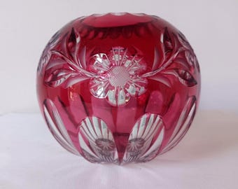 May include: A spherical ruby red cut-to-clear crystal vase featuring intricate floral engravings and vertical fluted patterns. The glass transitions from a deep red hue to clear sections at the base, showcasing detailed petal and leaf motifs.