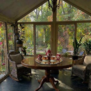 May include: A round wooden dining table with a pedestal base, featuring a mirrored tray holding a red and white porcelain tea set with a teapot, creamer, and two cups. Two wicker rocking chairs with beige cushions are placed on either side.