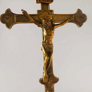 May include: A metallic gold-toned crucifix featuring a detailed figure of Jesus Christ centered on the cross. The cross has textured, darkened recessed areas and ornate, flared ends. The letters INRI are visible on a small plaque at the top.
