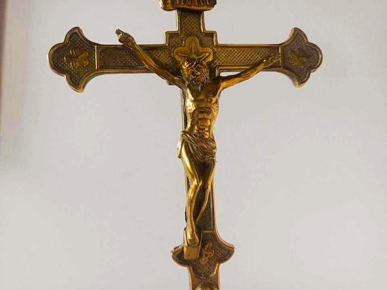 May include: A metallic gold-toned crucifix featuring a detailed figure of Jesus Christ centered on the cross. The cross has textured, darkened recessed areas and ornate, flared ends. The letters INRI are visible on a small plaque at the top.