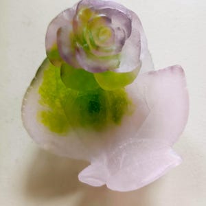 May include: A translucent, carved soap sculpture shaped like a blooming rose flower. The petals are a soft, pale pink with a gradient of vibrant green at the base, creating a layered, organic appearance with smooth, rounded edges and a glossy finish.